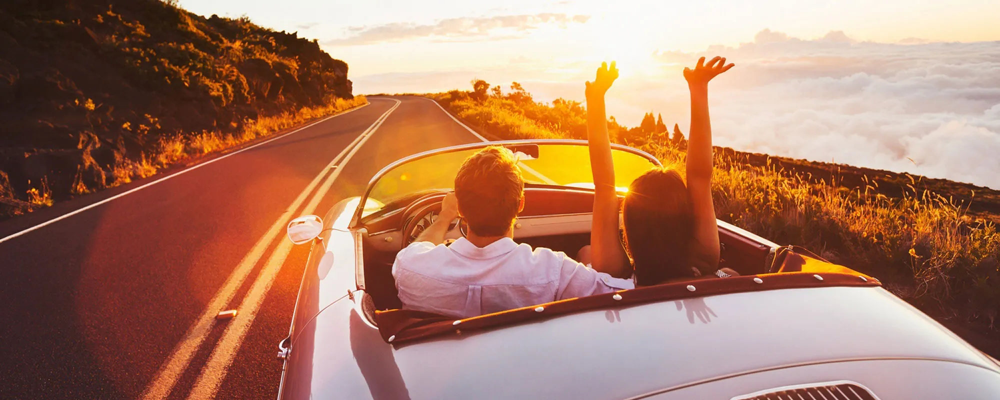 Couple driving a convertible car on a scenic road during a vibrant sunset.
