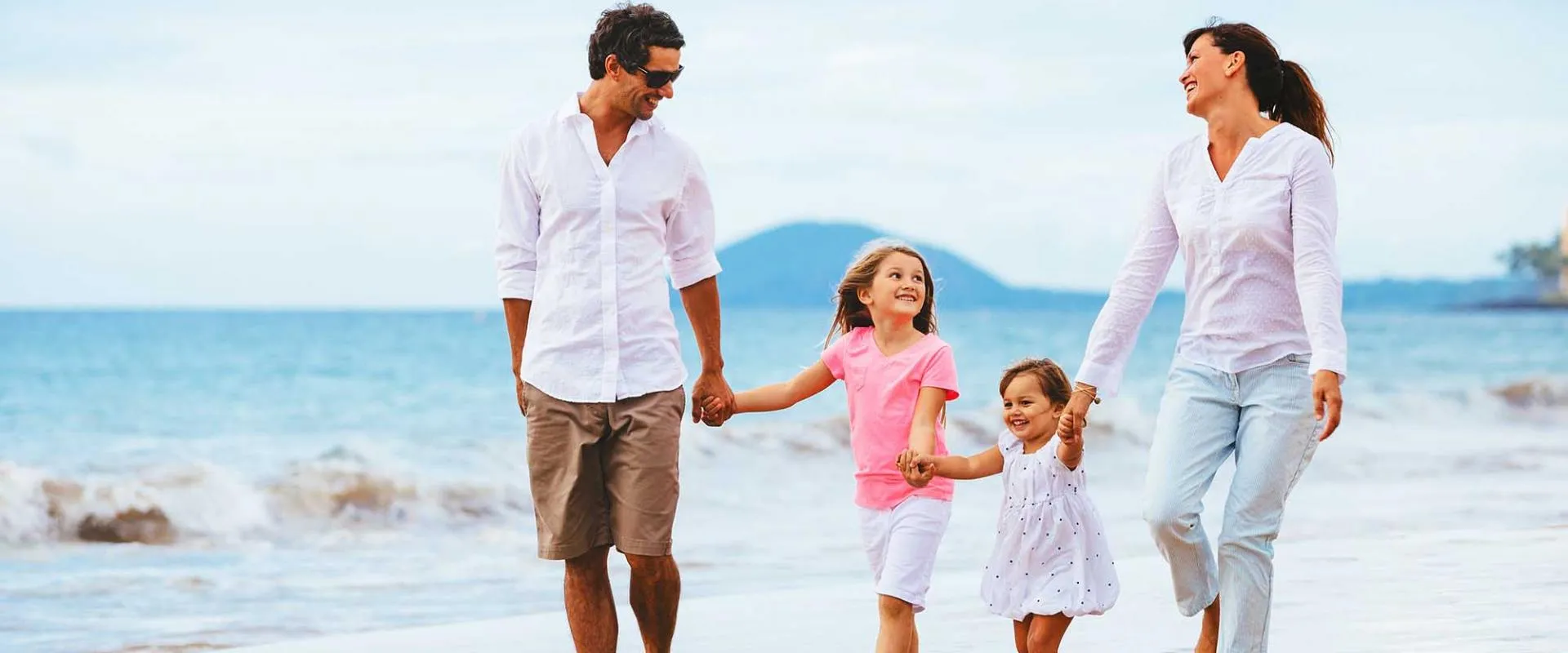 A family of four walks hand in hand along the beach, with waves gently lapping at the shore in the background.
